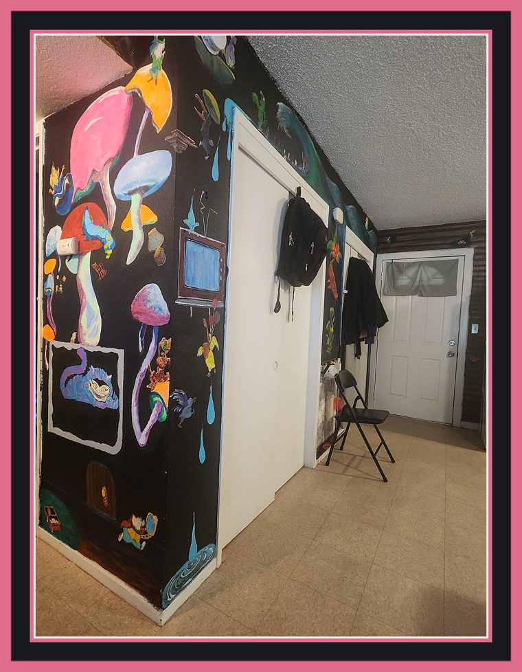 Angled hallway view of a black-ground mural of oversized psychedelic mushrooms in pink, blue and violet, with a small painted television, falling teardrops, and a framed Cheshire Cat panel painted by the artist's daughter.