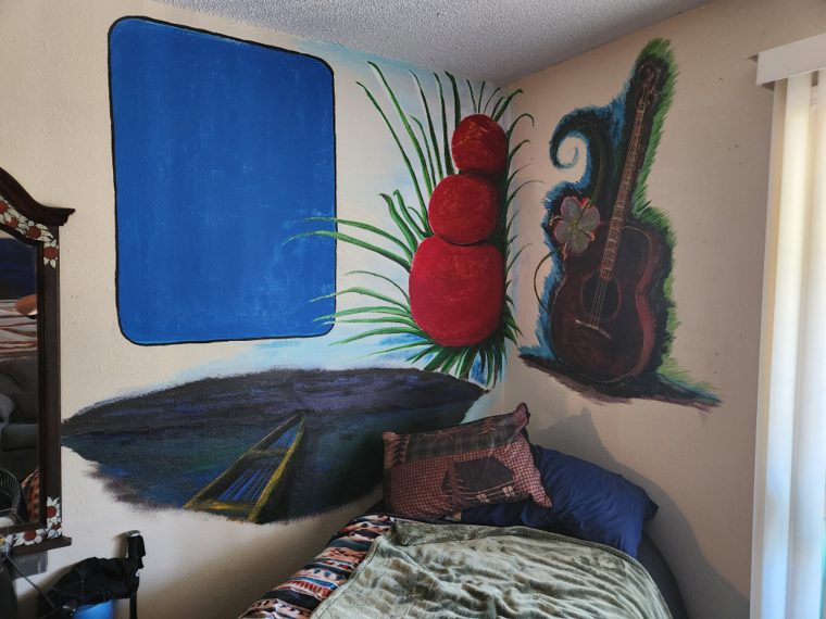 The corner mural with a new element added at the upper left: a flat blue rectangle blocked in with a black outline, the first pass at a painted styrofoam food package. The stacked red fruit, the grass, the dark landmass with its road, and the guitar in blue-green foliage are all further along.