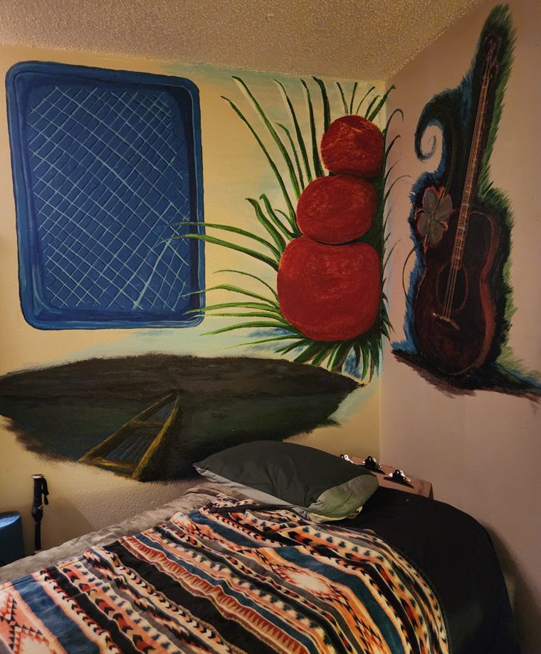 Latest state of the corner mural under warm light: the painted food package deepened and modelled at the upper left, the wall ground carried to a golden tone, the stacked red fruit and grass finished into it, and the dark landmass and its road resolved below.