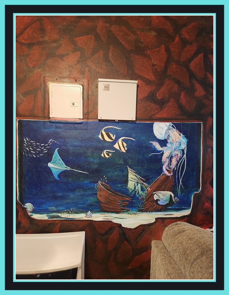 Painted aquarium set into an apartment wall of red-brown painted rock: a manta ray, two moorish idol fish, a drifting jellyfish, and a sunken shipwreck on a pale seabed.